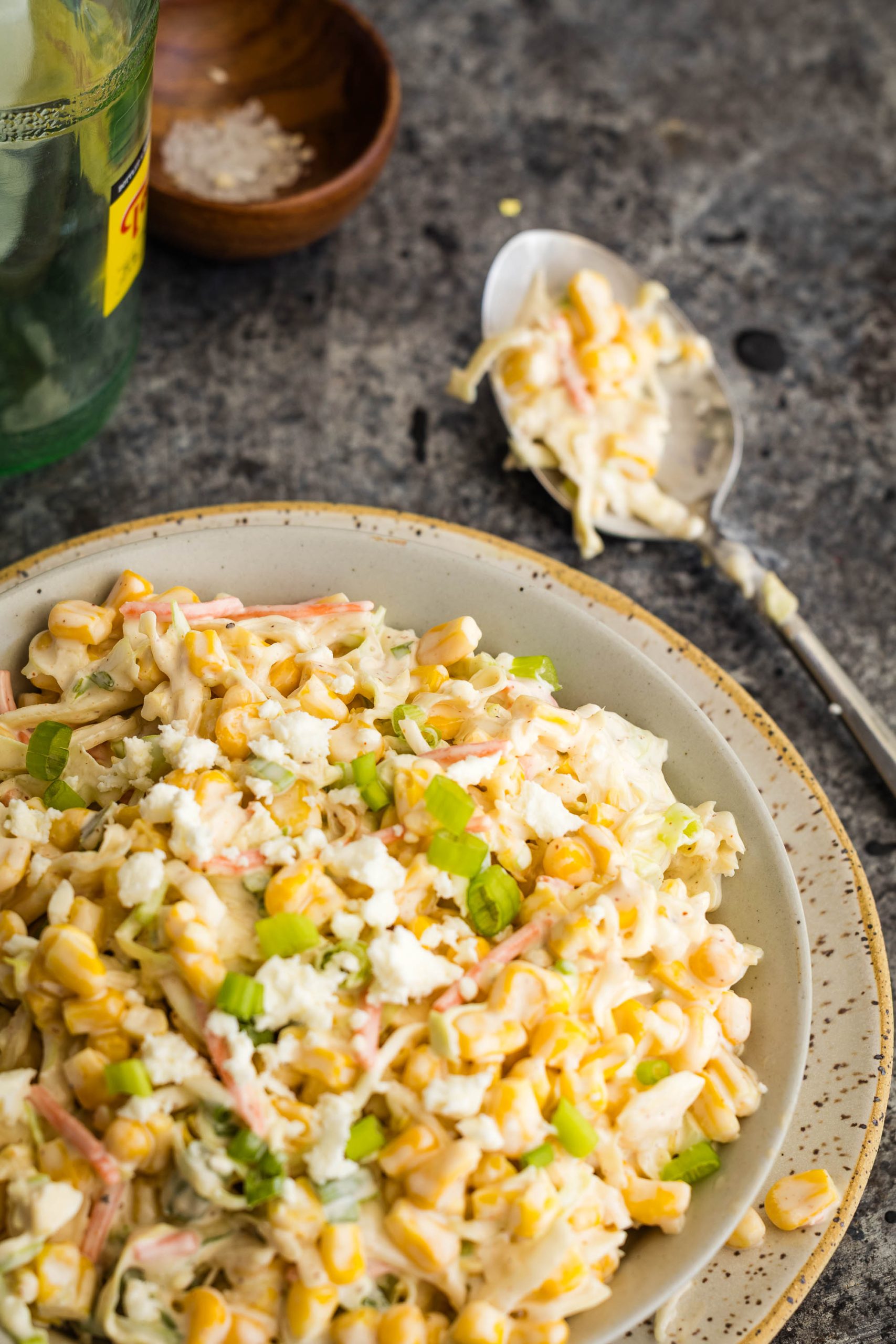 A bowl of corn salad with shredded cheese, carrots, and green onions sits on a rustic surface next to a spoonful of salad and a small bowl of salt.