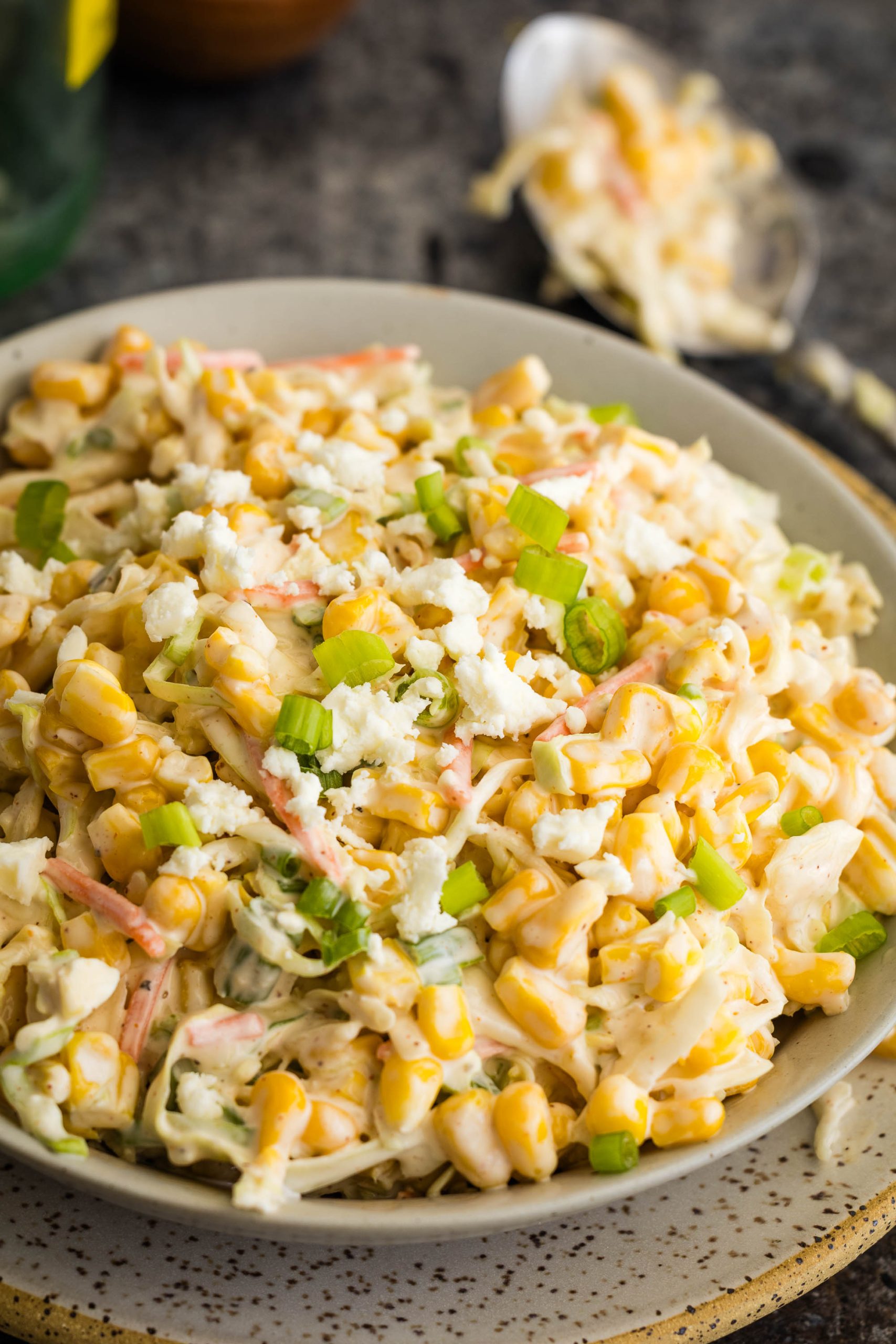 A plate of creamy corn salad topped with crumbled cheese and sliced green onions, with a spoon and more salad in the background.