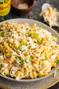 A plate of creamy corn salad topped with crumbled cheese and chopped green onions, with a bottle of Topo Chico and a small bowl of salt in the background.