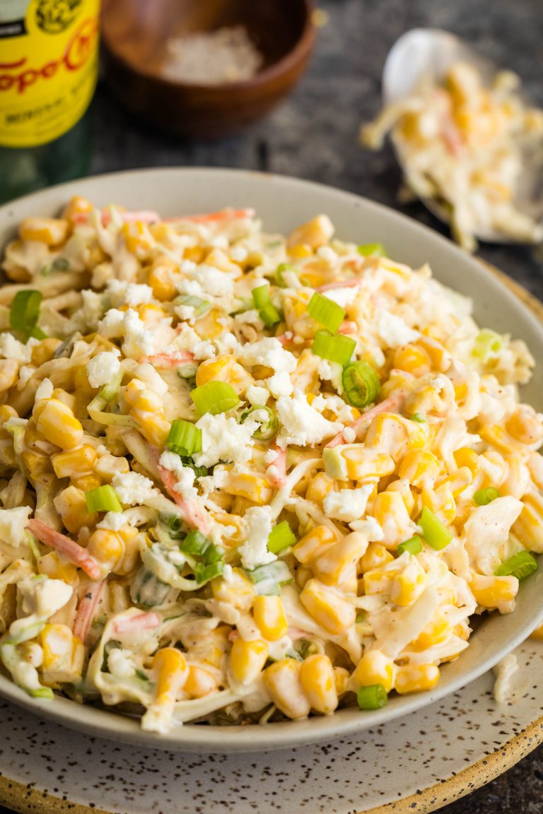 A plate of creamy corn salad topped with crumbled cheese and chopped green onions, with a bottle of Topo Chico and a small bowl of salt in the background.