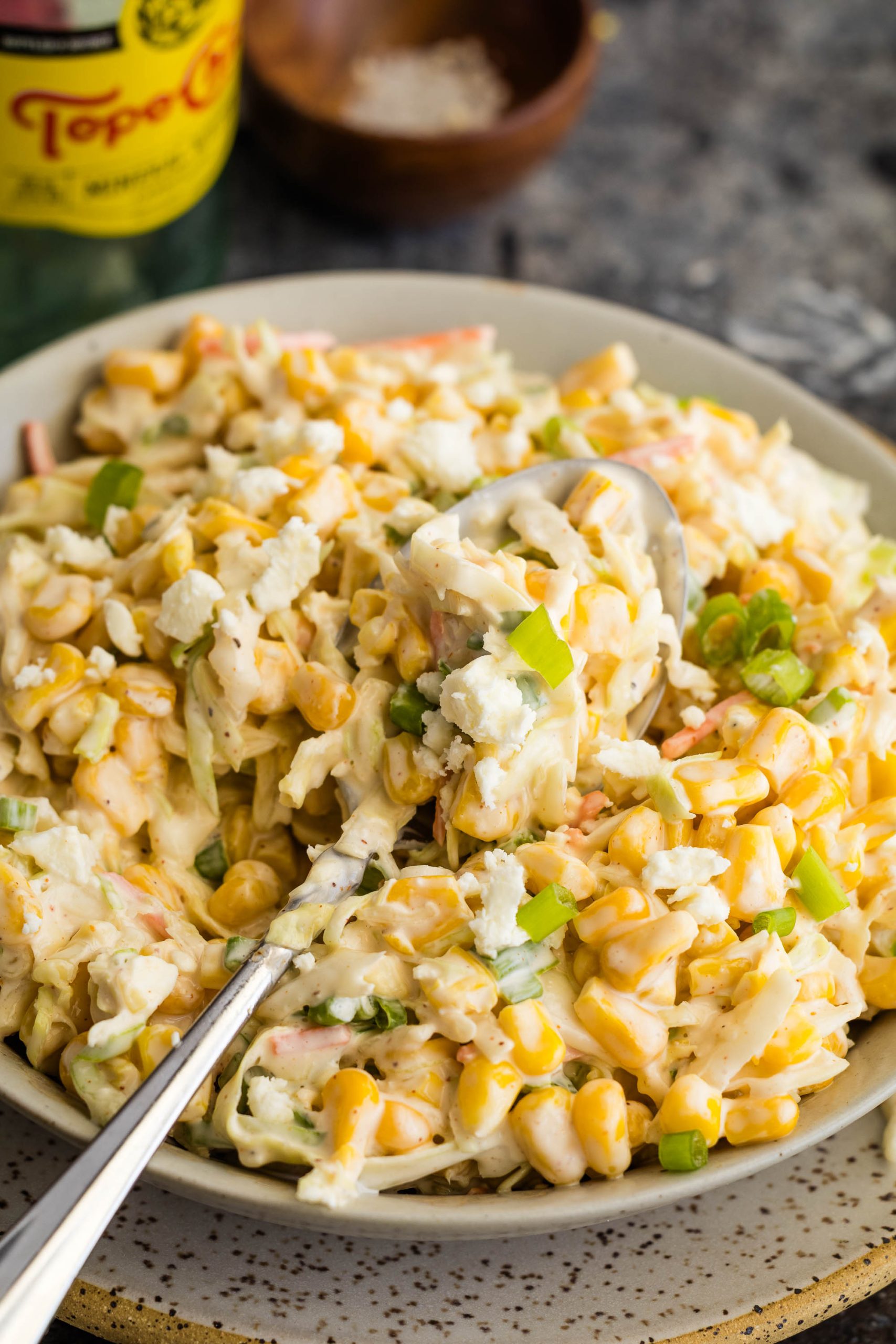 A close-up of a plate of creamy corn salad with crumbled cheese, chopped green onions, and a serving spoon.