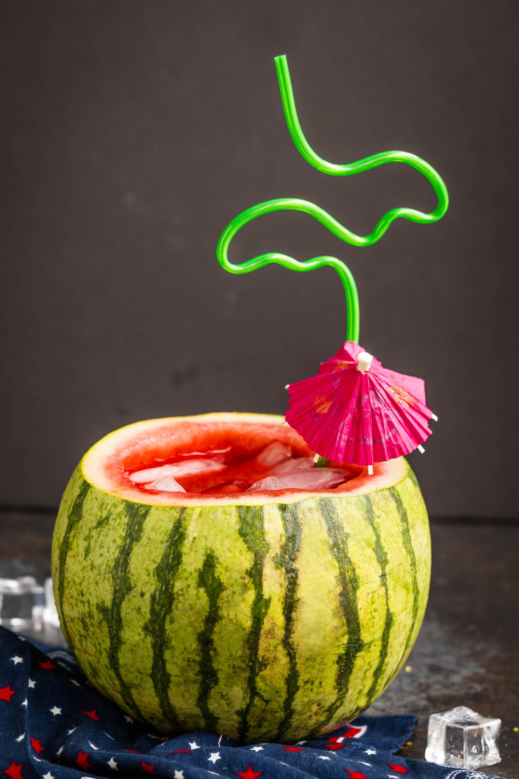 A hollowed-out watermelon filled with a refreshing watermelon cucumber mojito, ice cubes, a pink paper umbrella, and a green curly straw, set against a dark background.