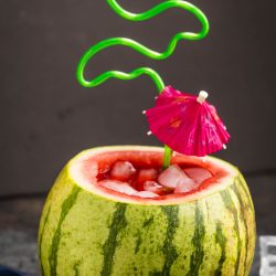 A hollowed-out watermelon filled with a refreshing watermelon cucumber mojito and ice cubes, topped with a green curly straw and a pink cocktail umbrella.