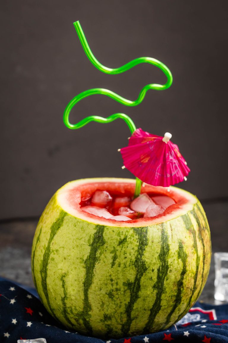 A hollowed-out watermelon filled with a refreshing watermelon cucumber mojito and ice cubes, topped with a green curly straw and a pink cocktail umbrella.