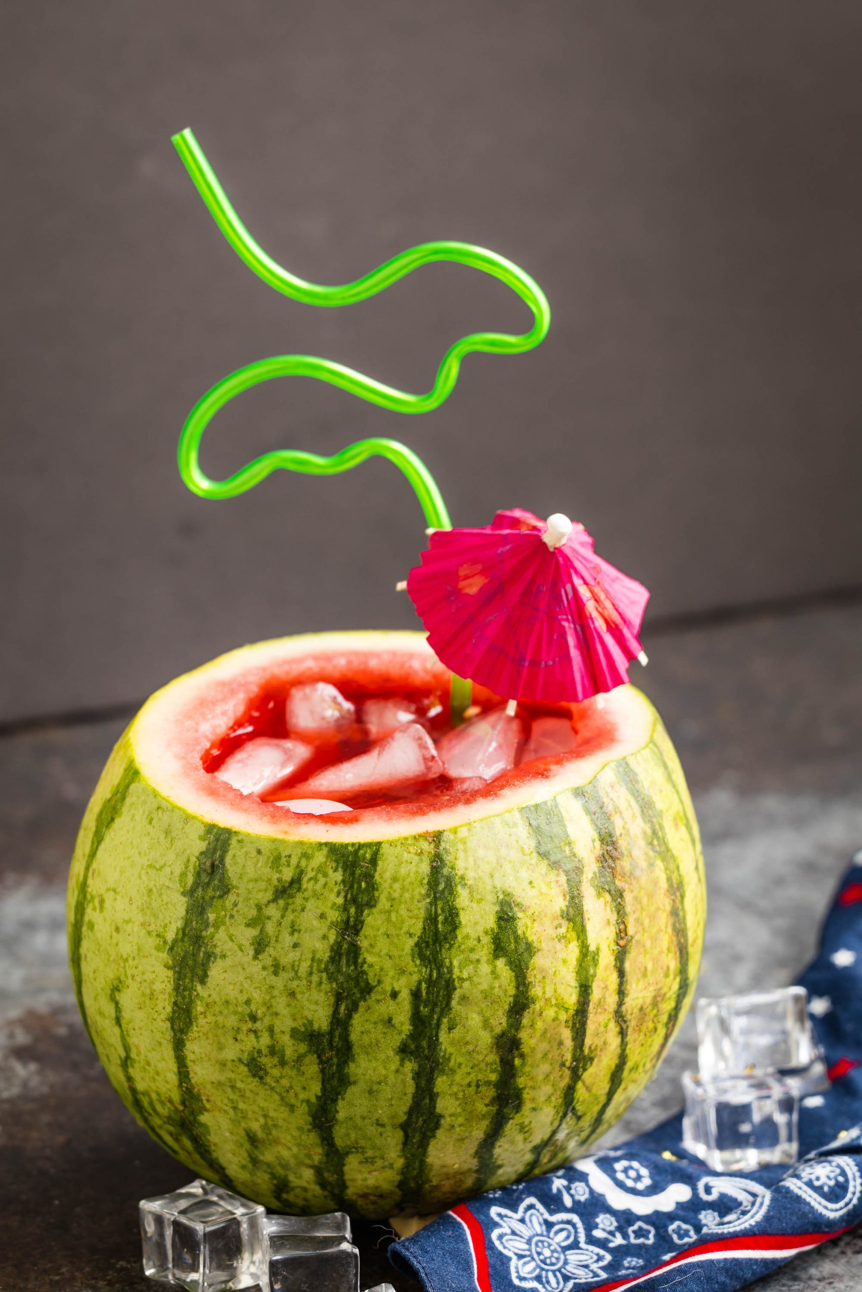 A hollowed-out watermelon filled with drink and ice cubes, garnished with a pink cocktail umbrella and a curly green straw.