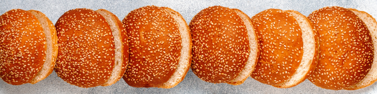 Six sesame seed hamburger buns, perfect for crafting the Best Burgers, are arranged in a horizontal line on a light gray surface.