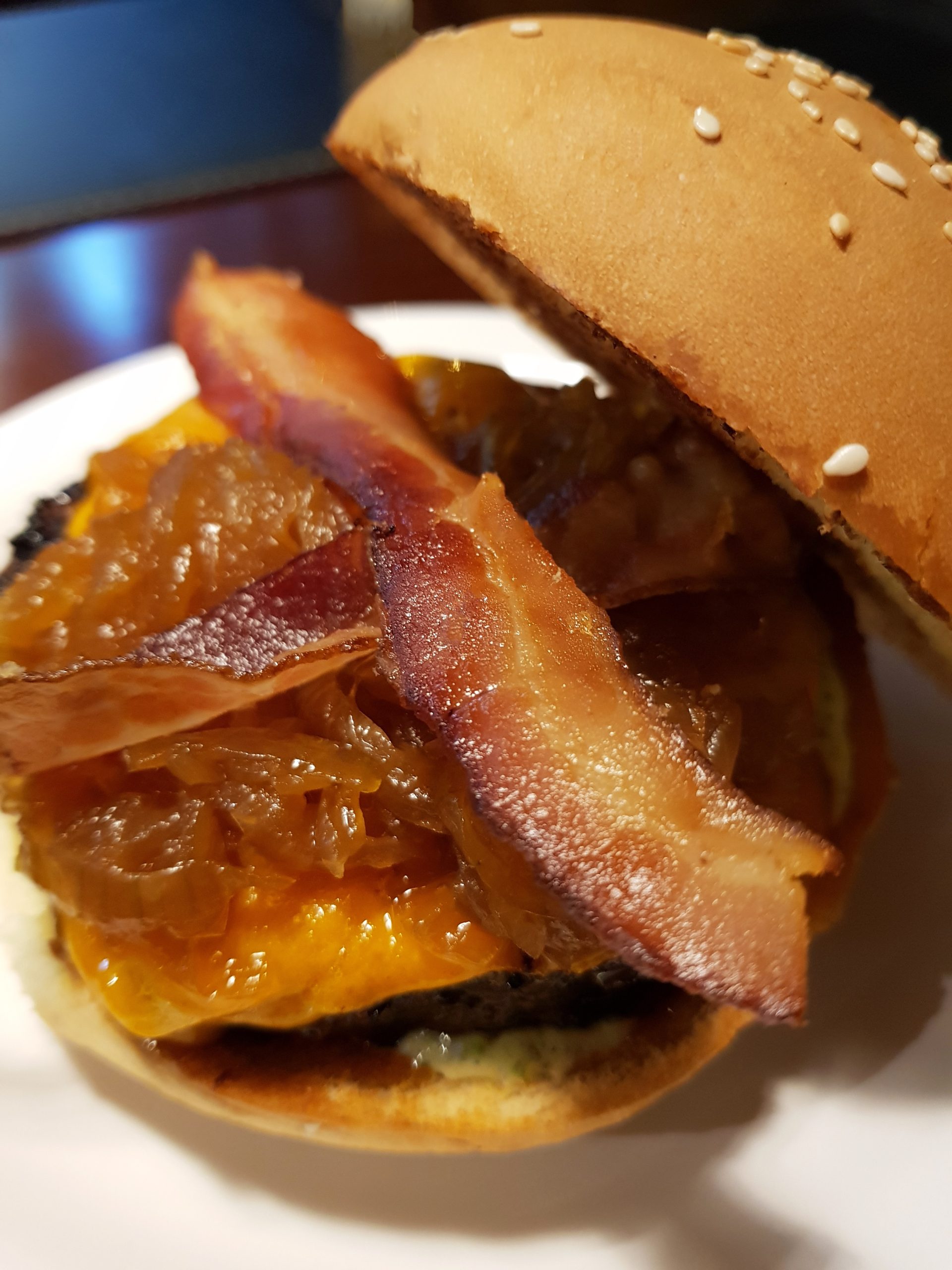 A close-up of food—featuring crispy bacon strips, caramelized onions, and a sesame seed bun—served on a white plate.