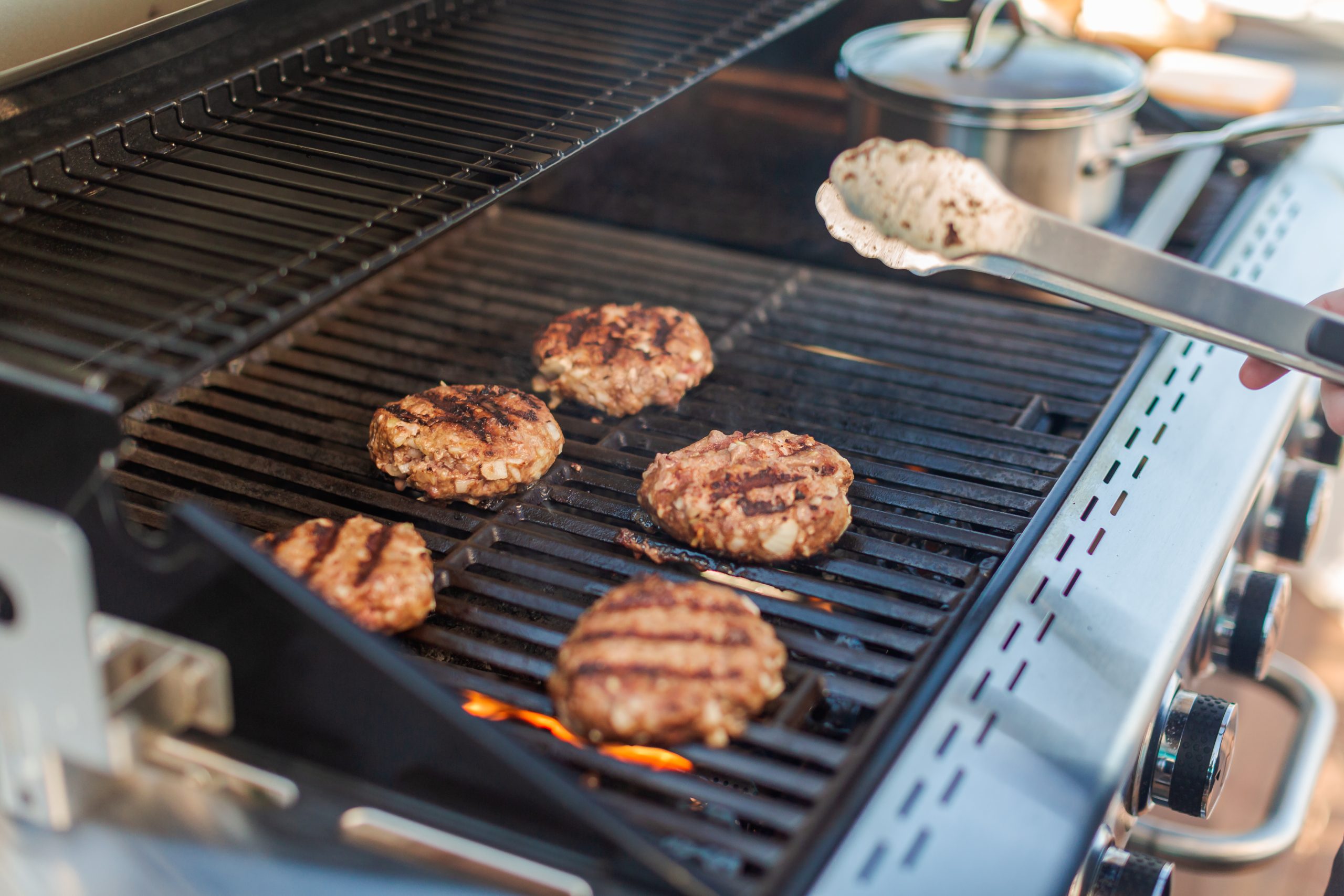 Five burger patties cooking on a gas grill; a hand with tongs holds another patty over the grill. A metal pot with a lid sits on the side burner.