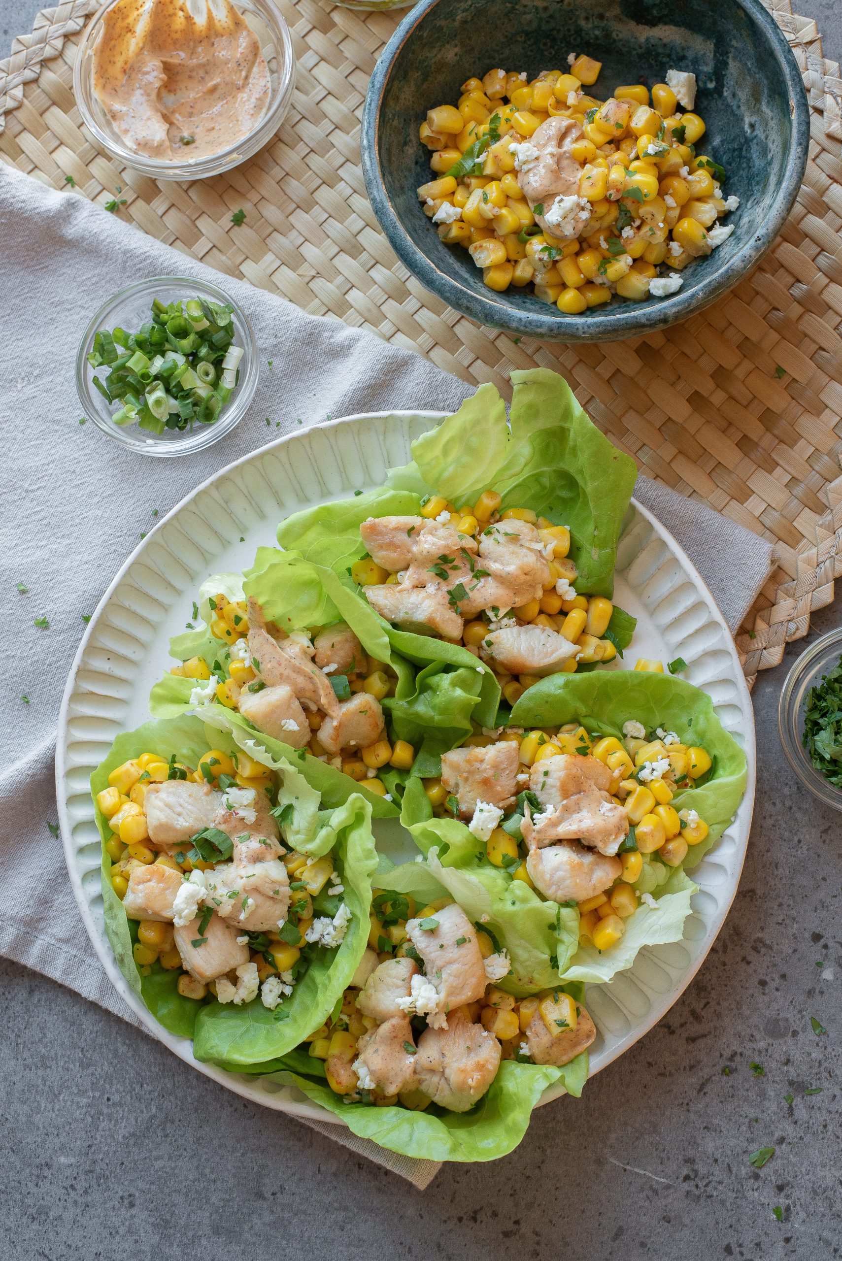 Three lettuce wraps filled with diced chicken, corn, cheese, and herbs on a plate, with bowls of corn, sauce, and green onions on the side.