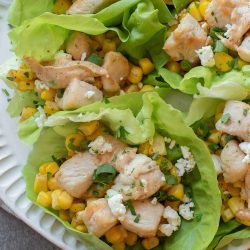 Four lettuce wraps filled with diced chicken, corn, and crumbled cheese are arranged on a white plate.