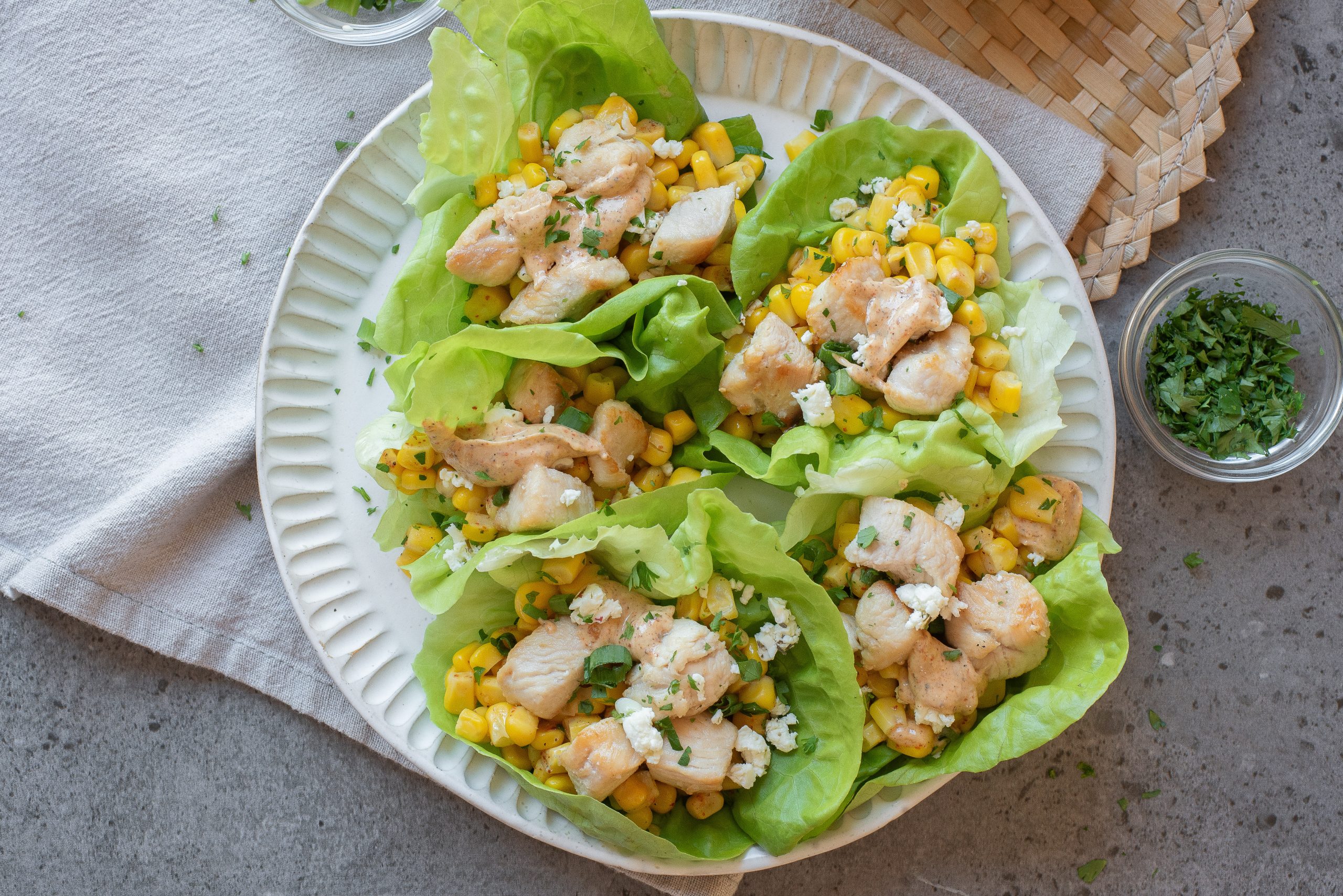 A plate with four Street Corn Chicken Lettuce Wraps filled with diced chicken, corn, chickpeas, and crumbled cheese, garnished with chopped herbs.