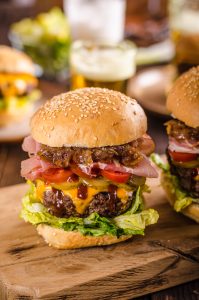 A hamburger with lettuce, cheddar cheese, tomato, onions, ham, and sauce in a sesame seed bun on a wooden board. Another burger is slightly visible beside it.