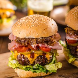A hamburger with lettuce, cheddar cheese, tomato, onions, ham, and sauce in a sesame seed bun on a wooden board. Another burger is slightly visible beside it.
