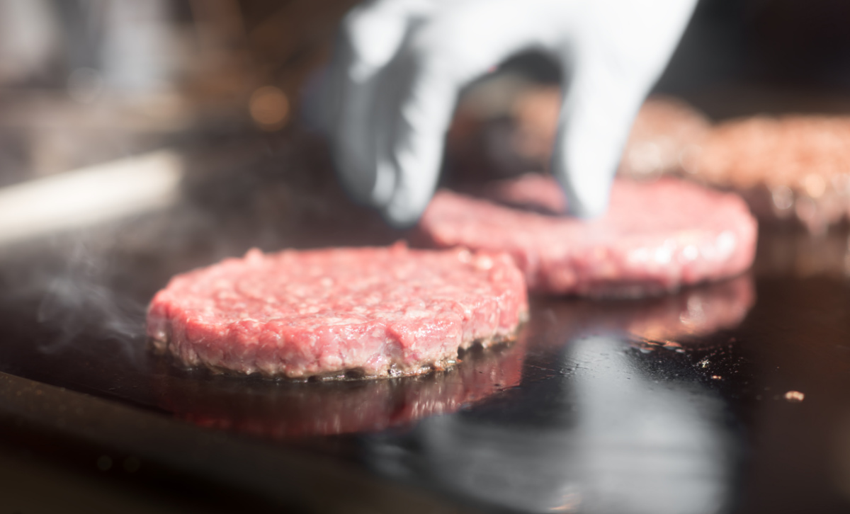 A gloved hand places raw beef patties on a hot grill, with steam rising as they cook.