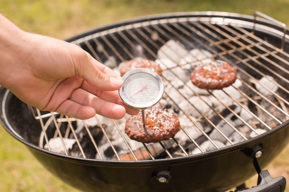 A hand holds a meat thermometer above burgers cooking on a charcoal grill, ensuring every patty reaches the perfect temperature for the Best Burgers.