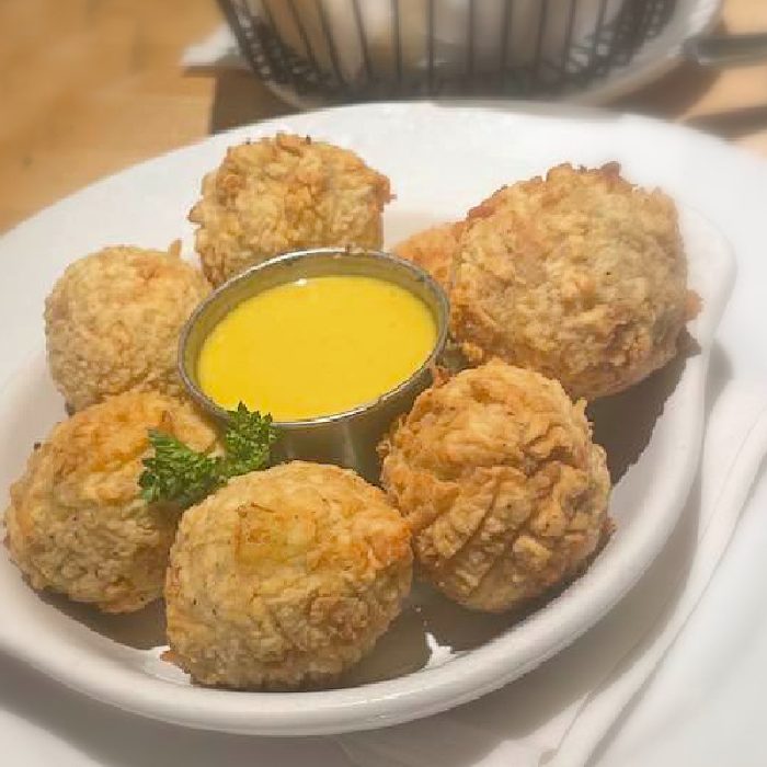 Six breaded and fried balls arranged around a cup of yellow dipping sauce on a white plate, garnished with a small sprig of parsley—an appetizing starter from Harvey's Tupelo MS.