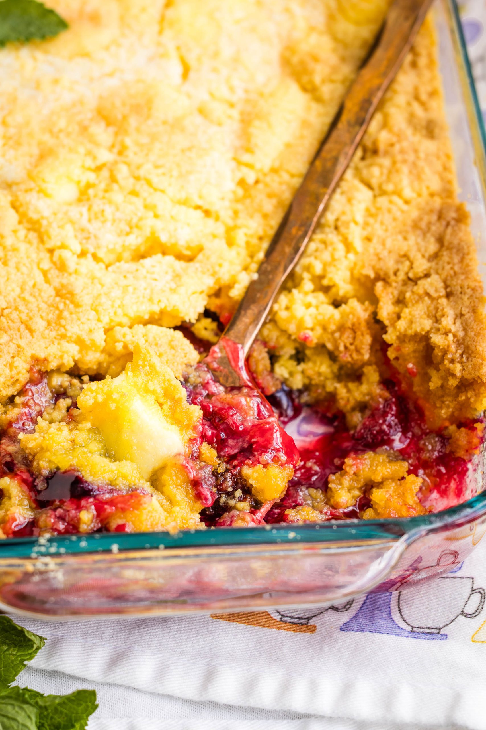 A close-up of a partially served Berry Cream Cheese Cobbler in a glass baking dish, with a serving spoon resting inside and a visible golden, crumbly topping.
