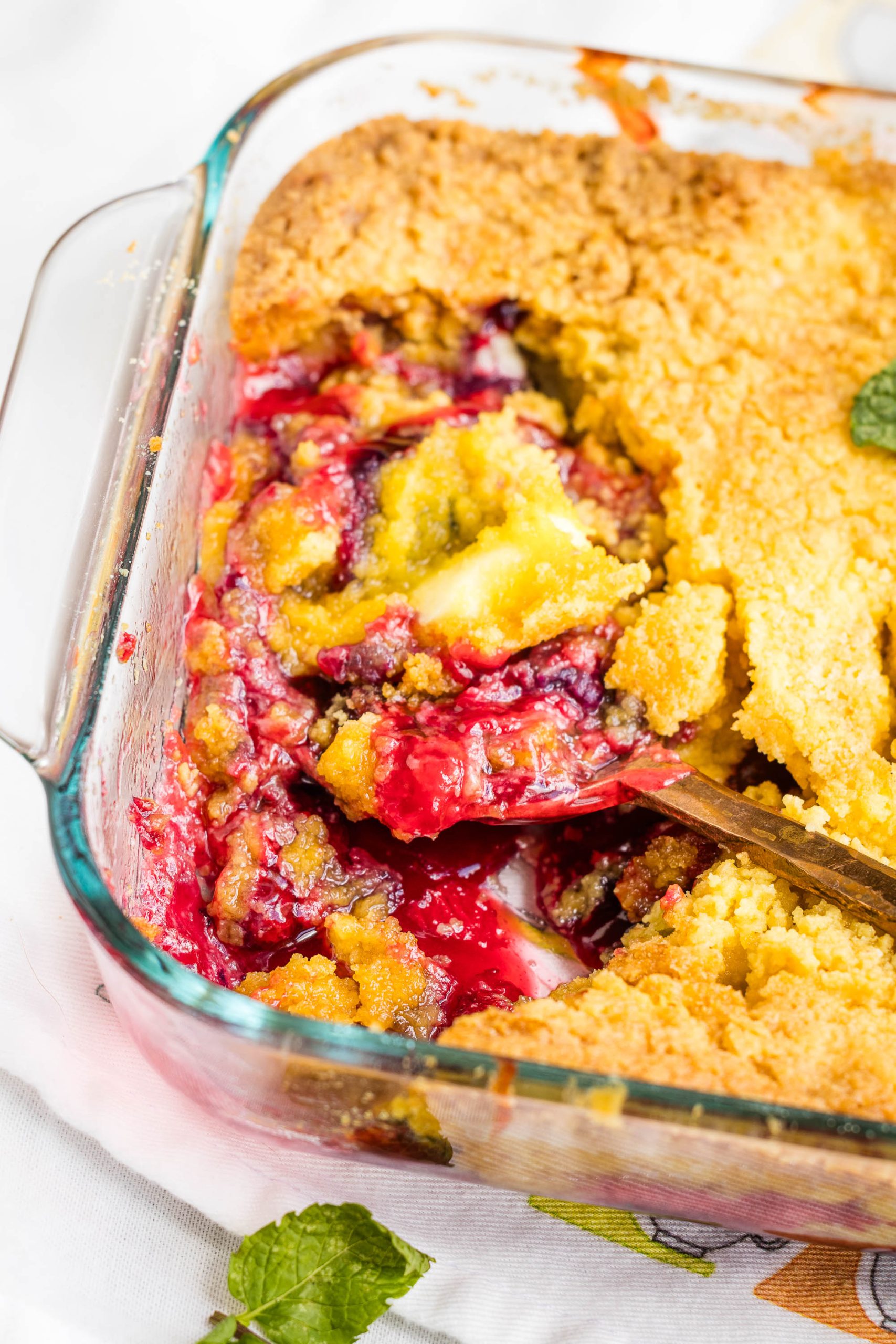 A glass baking dish with a partially served Berry Cream Cheese Cobbler, showing golden crust and red fruit filling, with a serving spoon inside.