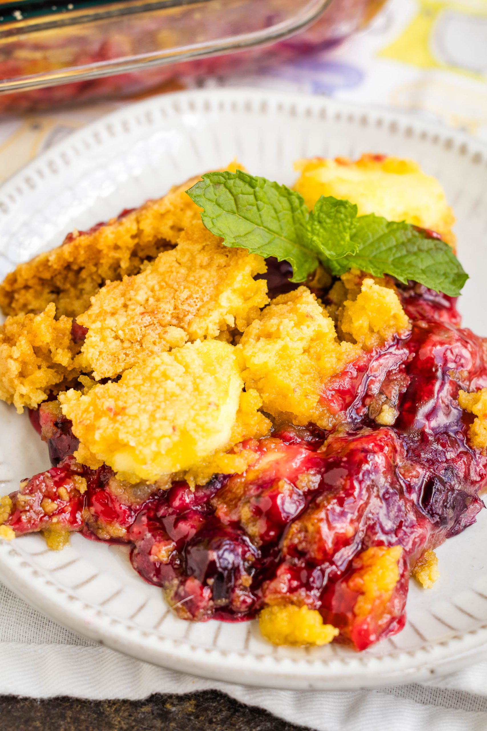 A serving of berry cobbler with a golden crumb topping, garnished with a sprig of fresh mint, on a white plate.