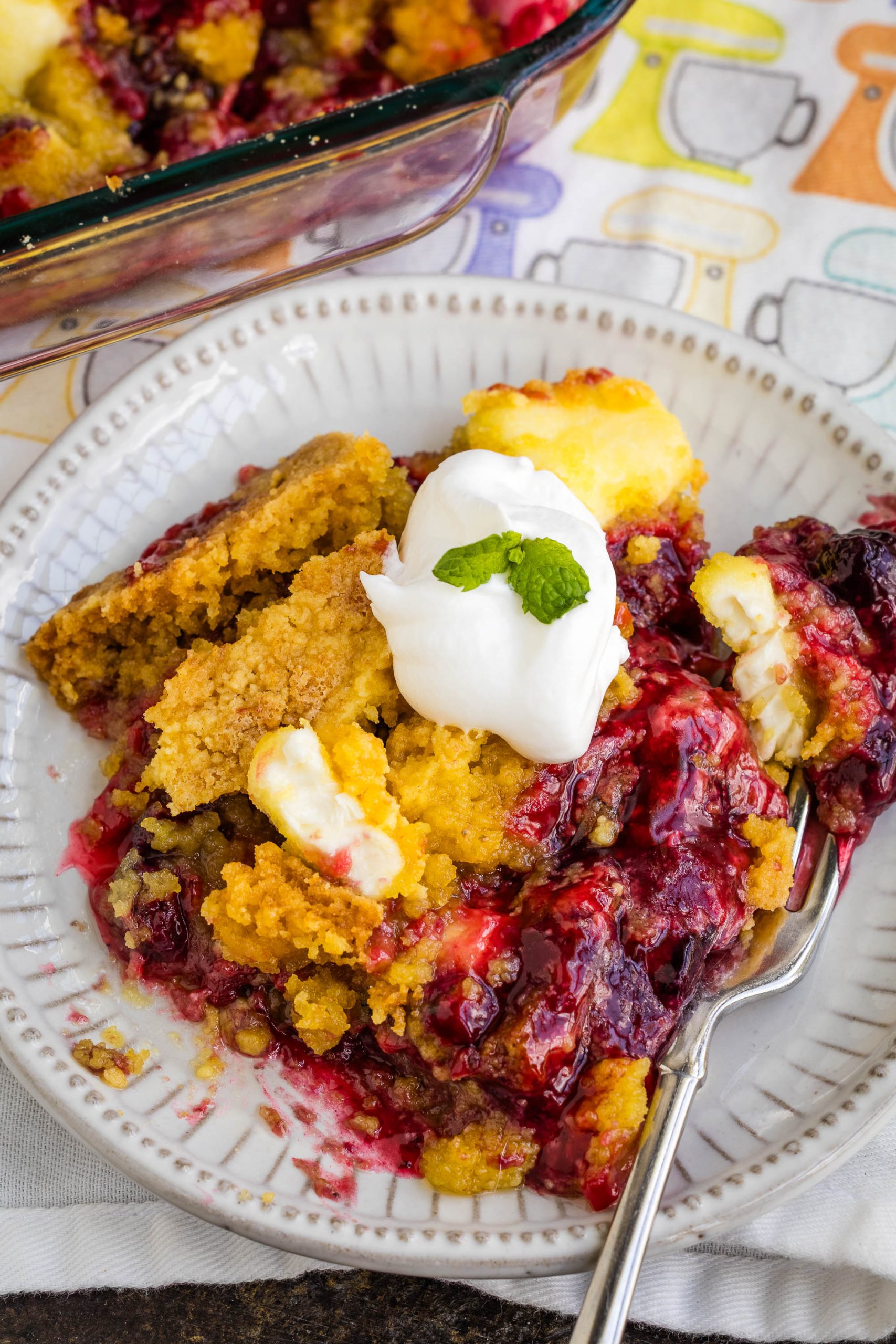 A plate of Berry Cream Cheese Cobbler topped with whipped cream and a mint leaf, with a fork, sits on a patterned tablecloth near a baking dish of more cobbler.