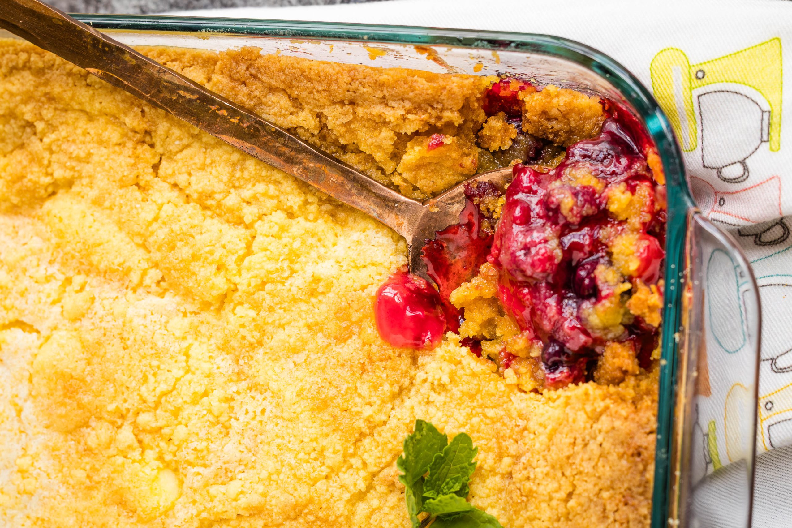 A close-up of a baked cherry crumble dessert in a glass dish with a serving spoon and a garnish of fresh mint.