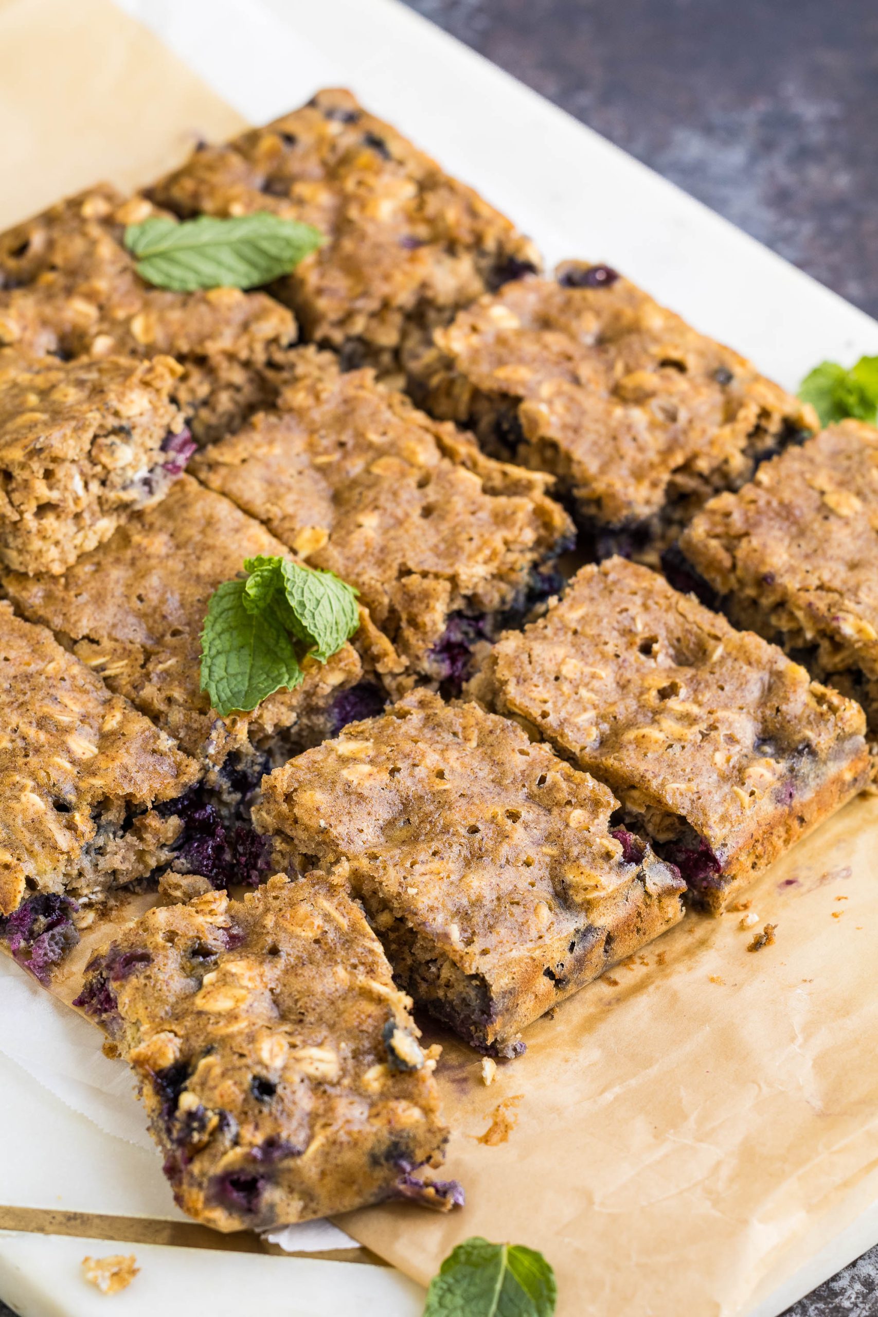 A batch of Blueberry Oatmeal Breakfast Blondies, sliced and arranged on parchment paper, features visible blueberries and is garnished with fresh mint leaves.