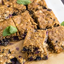 Nine Blueberry Oatmeal Breakfast Blondies are arranged on parchment paper, with a few bars stacked and garnished with fresh mint leaves.