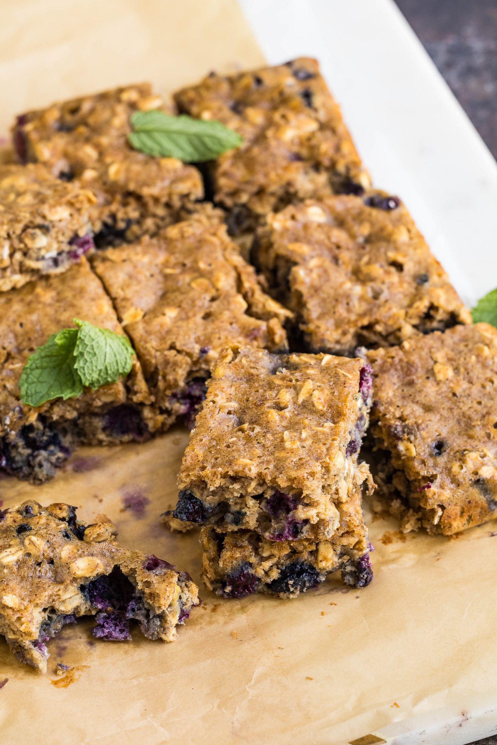 Nine Blueberry Oatmeal Breakfast Blondies are arranged on parchment paper, with a few bars stacked and garnished with fresh mint leaves.