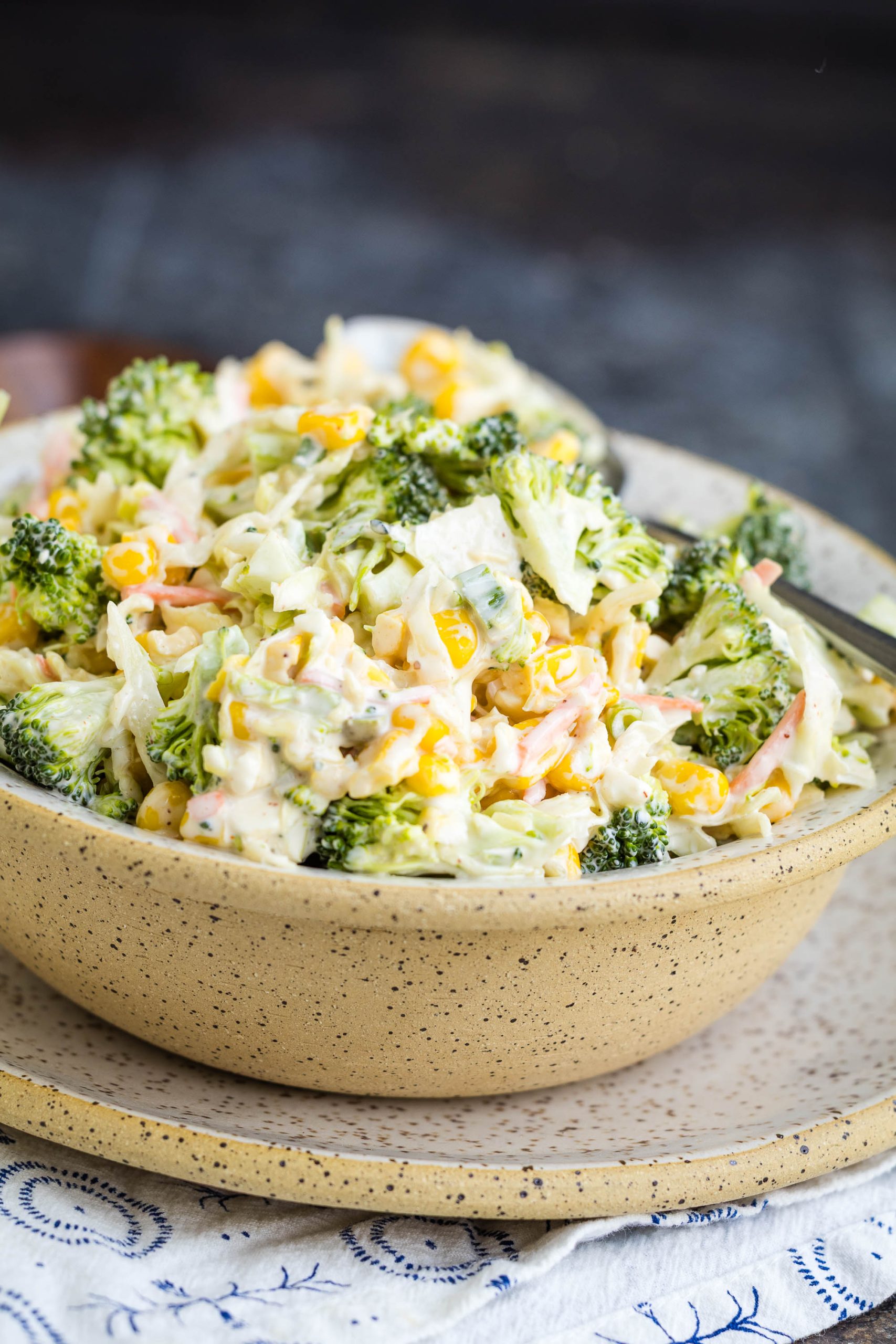 A bowl of creamy broccoli salad with shredded cheese, carrots, and mayonnaise, served in a speckled ceramic bowl on a matching plate.