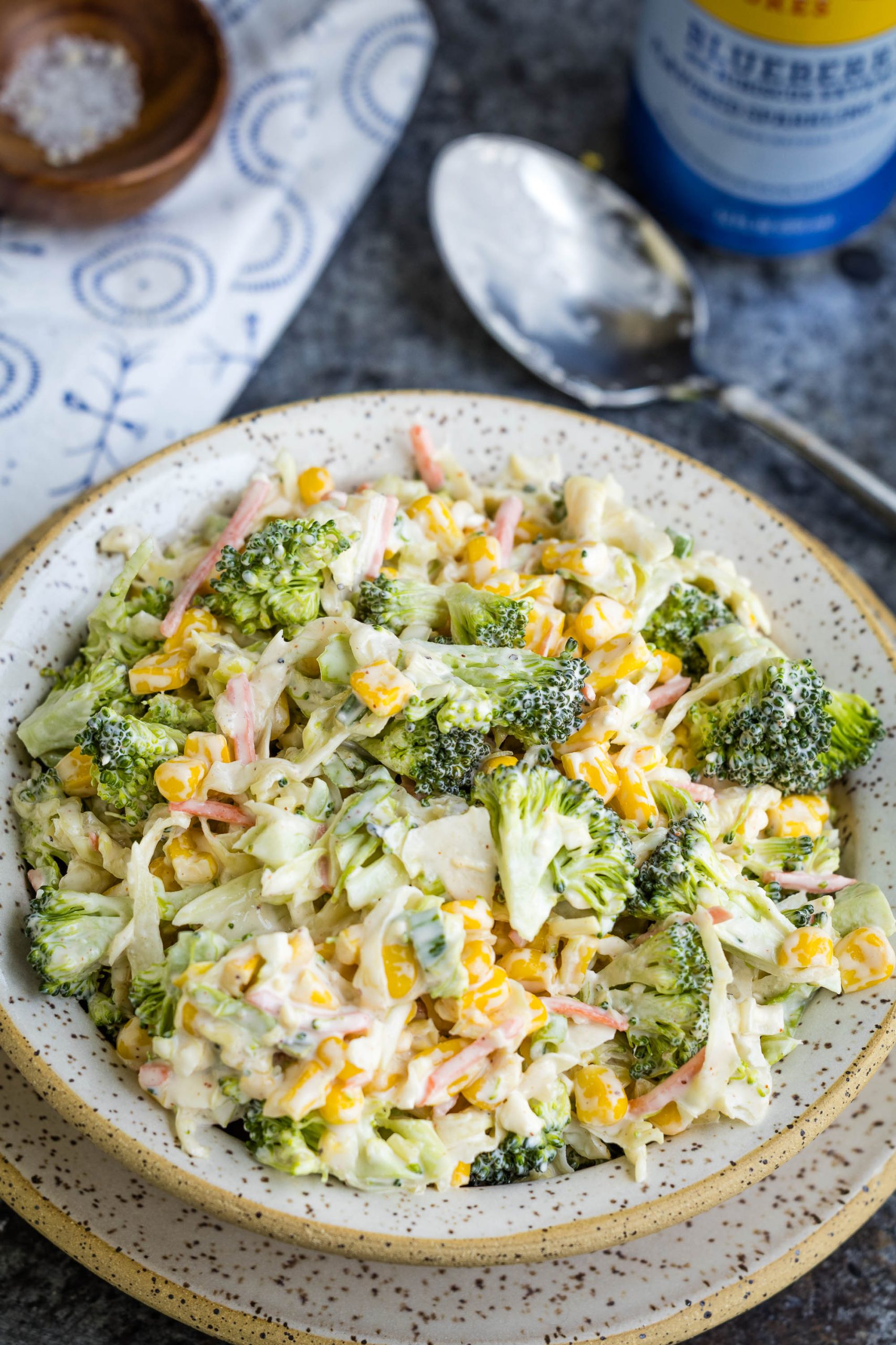 A bowl of broccoli salad mixed with shredded carrots, corn, and a creamy dressing, placed on a plate with a spoon and salt dish nearby.