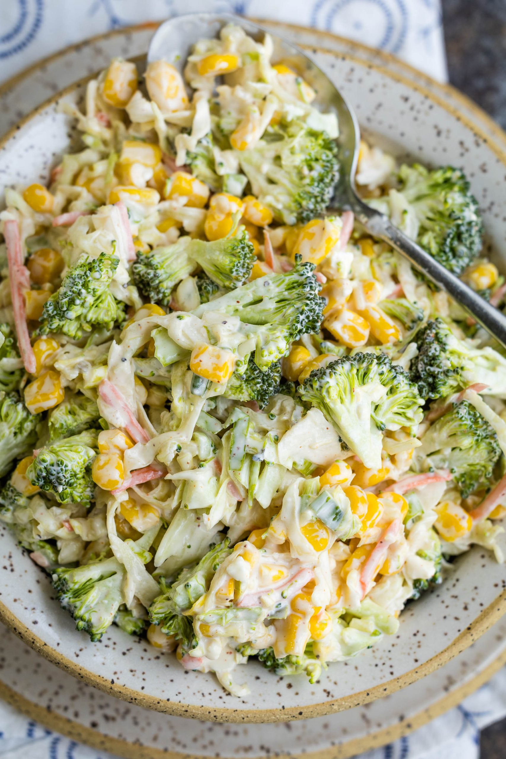 A plate of broccoli salad with corn, shredded vegetables, and a creamy dressing, served with a spoon.