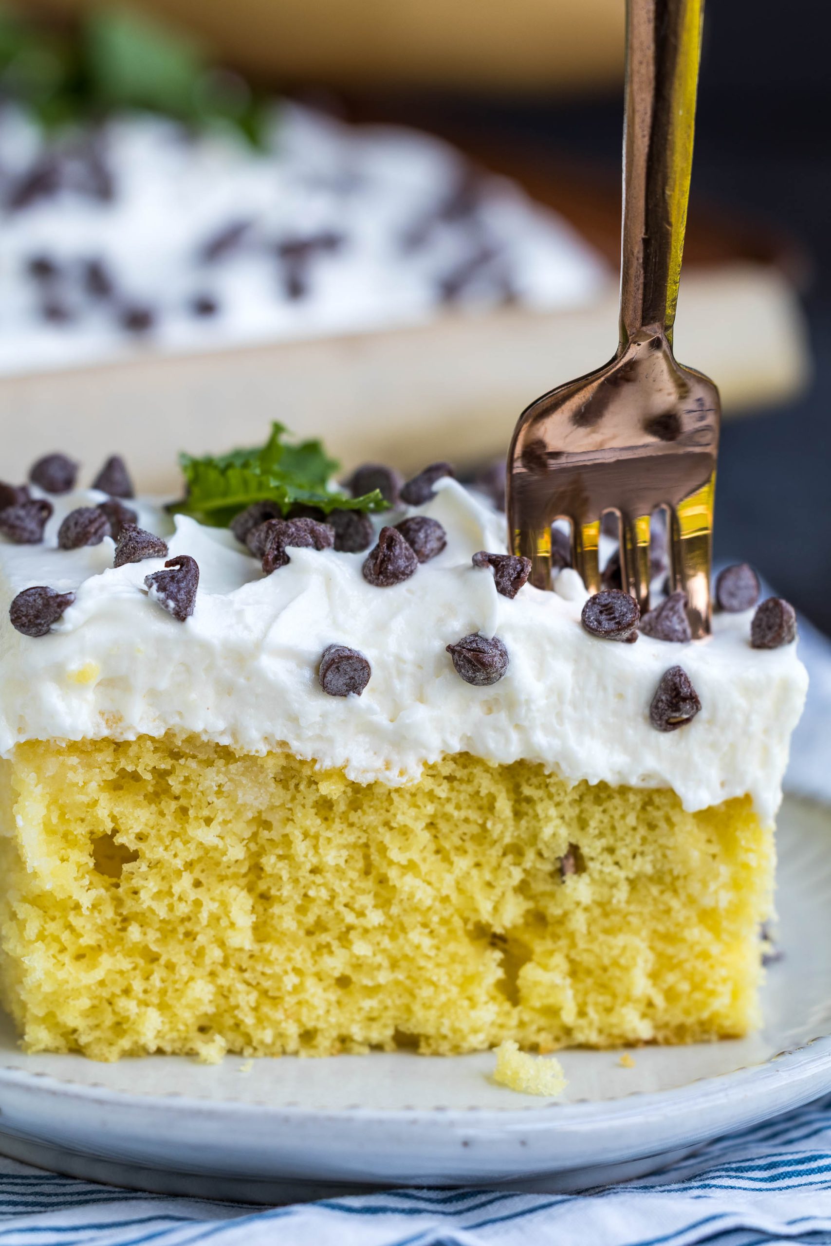 A fork is inserted into a slice of cannoli sheet cake topped with whipped cream and chocolate chips, served on a white plate.