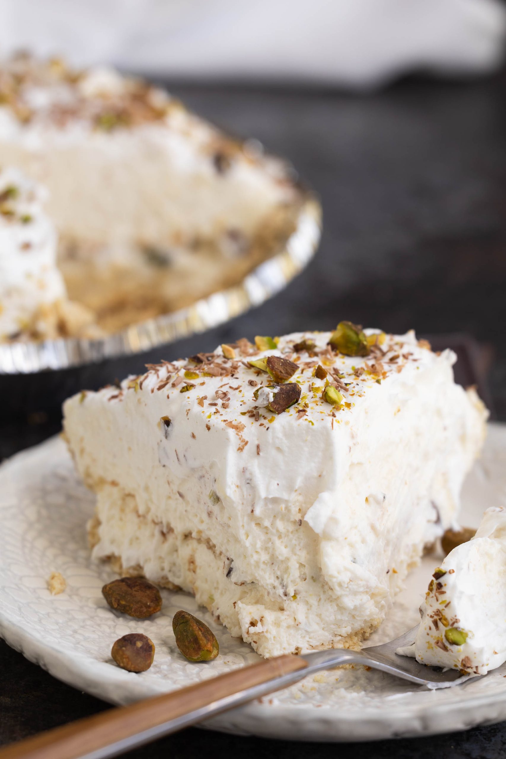 A slice of creamy pie topped with whipped cream, chocolate shavings, and pistachios on a white plate, with the rest of the pie in the background.