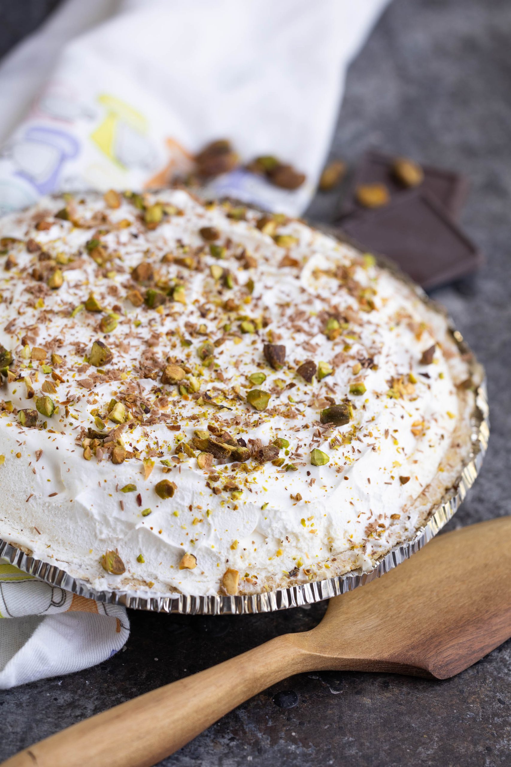 A cream pie topped with chopped pistachios and chocolate shavings in a foil pie tin, with a wooden spatula beside it on a dark countertop.