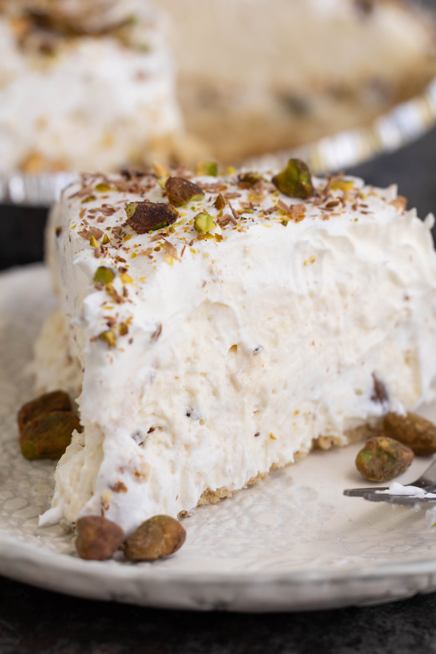 A slice of creamy pie topped with chopped nuts sits on a white plate, with a fork and additional nuts beside it.