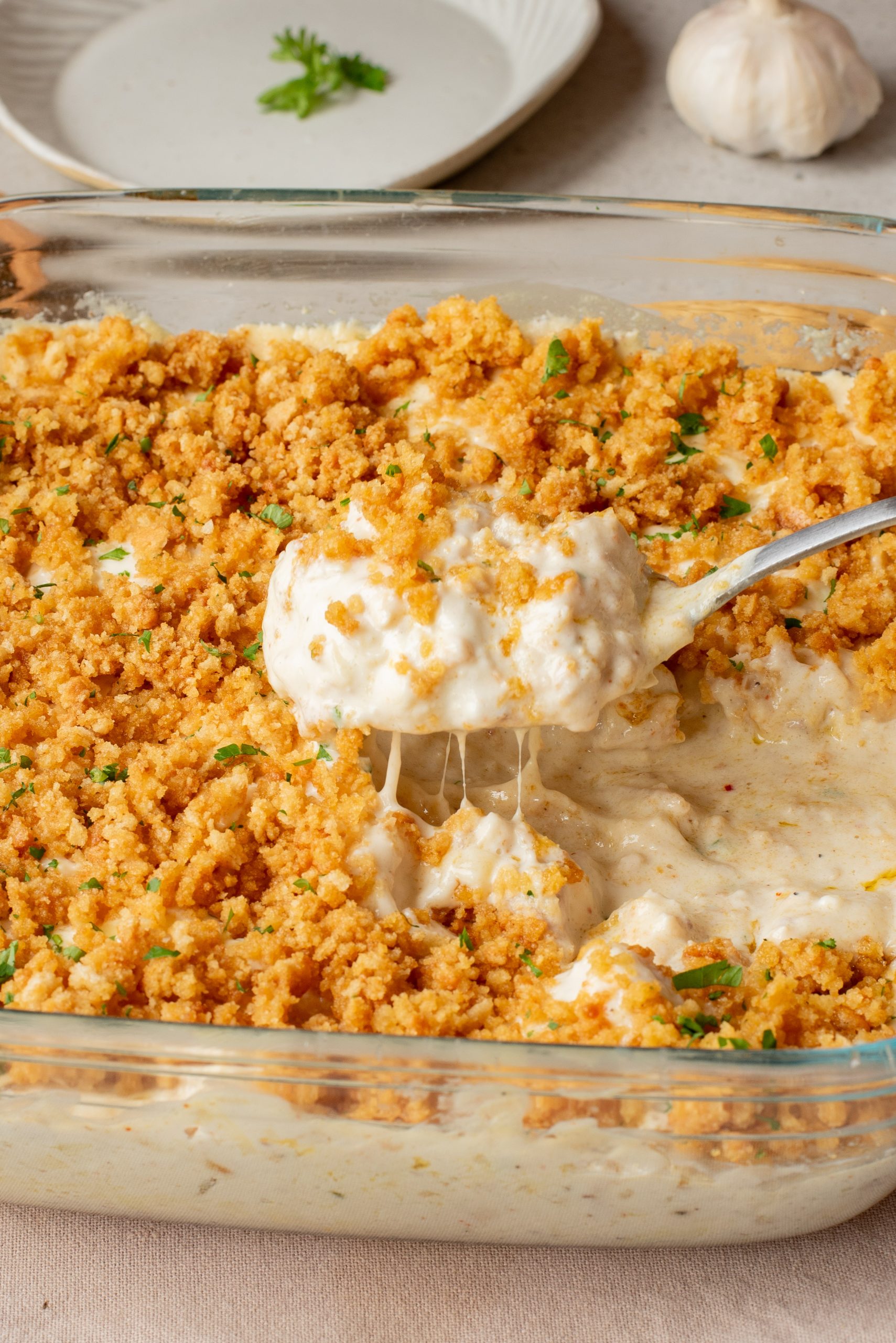 A glass baking dish filled with a cheesy, creamy casserole topped with golden breadcrumbs; a spoon is lifting out a serving.