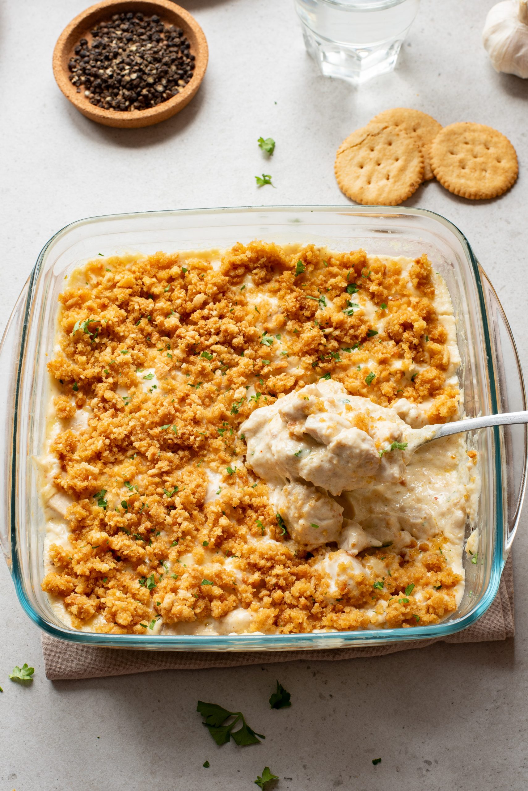A glass baking dish filled with creamy casserole topped with golden breadcrumbs, with a serving spoon scooping some out. Crackers, pepper, and a glass of water are nearby.