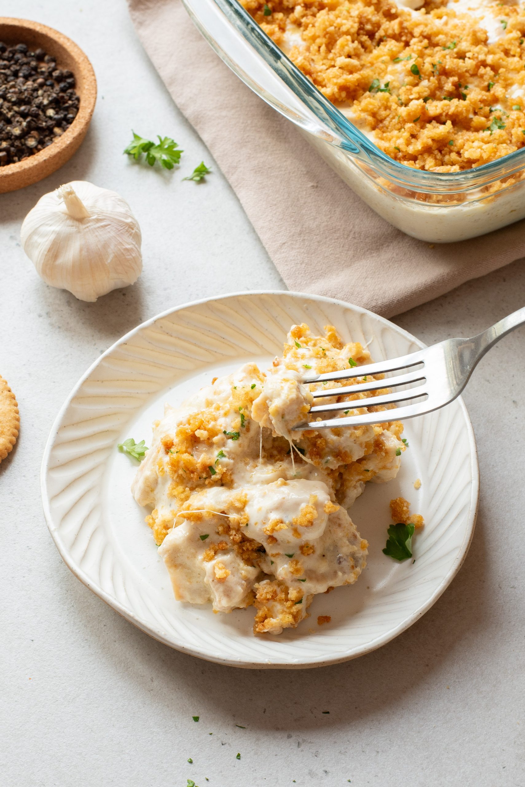 A serving of creamy casserole with a golden breadcrumb topping on a white plate, being picked up with a fork; casserole dish, garlic, and pepper visible nearby.