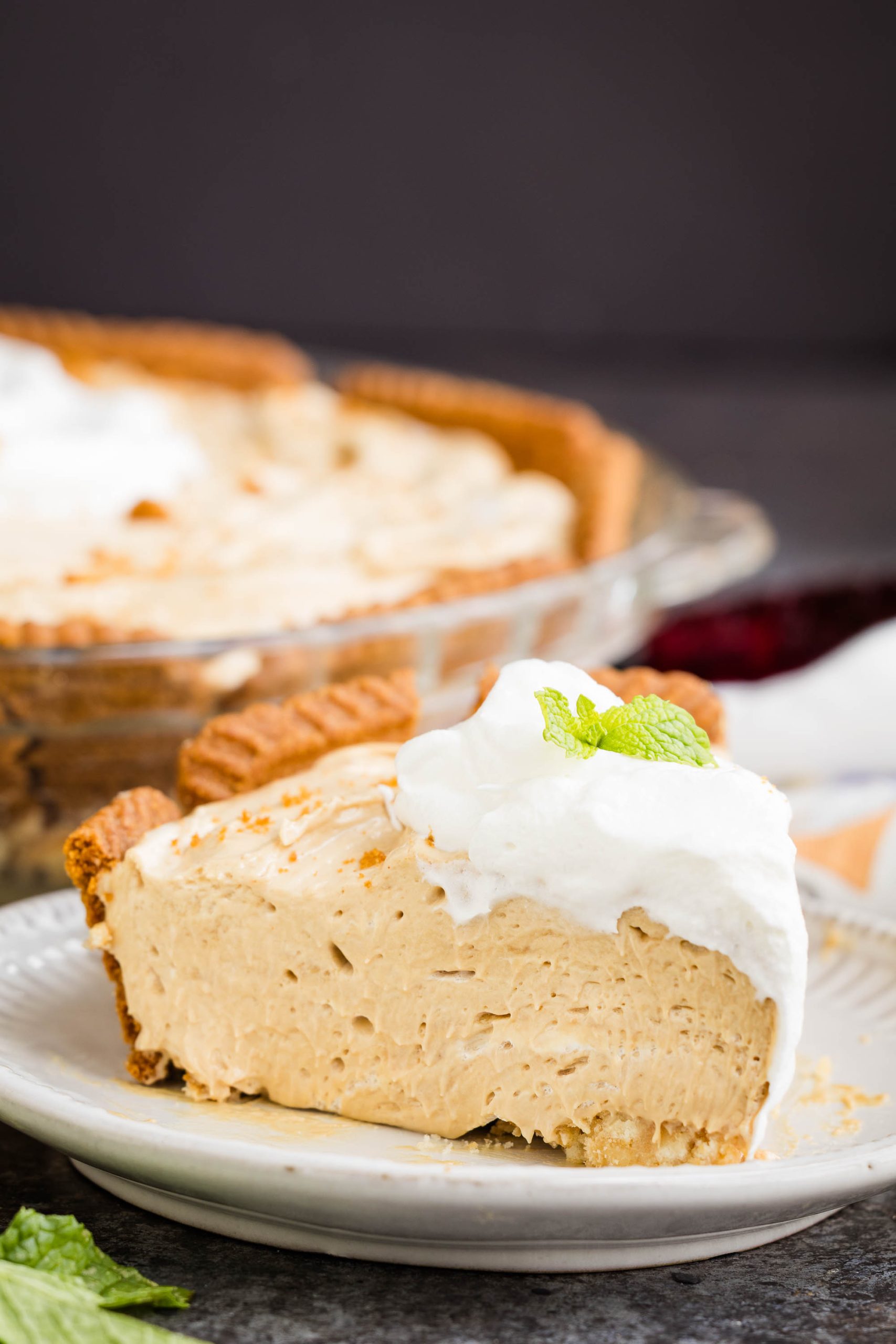 A slice of creamy pie with whipped topping and a mint leaf, served on a plate, with the rest of the pie in a glass dish in the background.