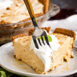 A fork holds a bite of creamy pie with whipped topping on a plate, with the rest of the pie in a glass dish in the background. Mint leaves are also visible on the table.
