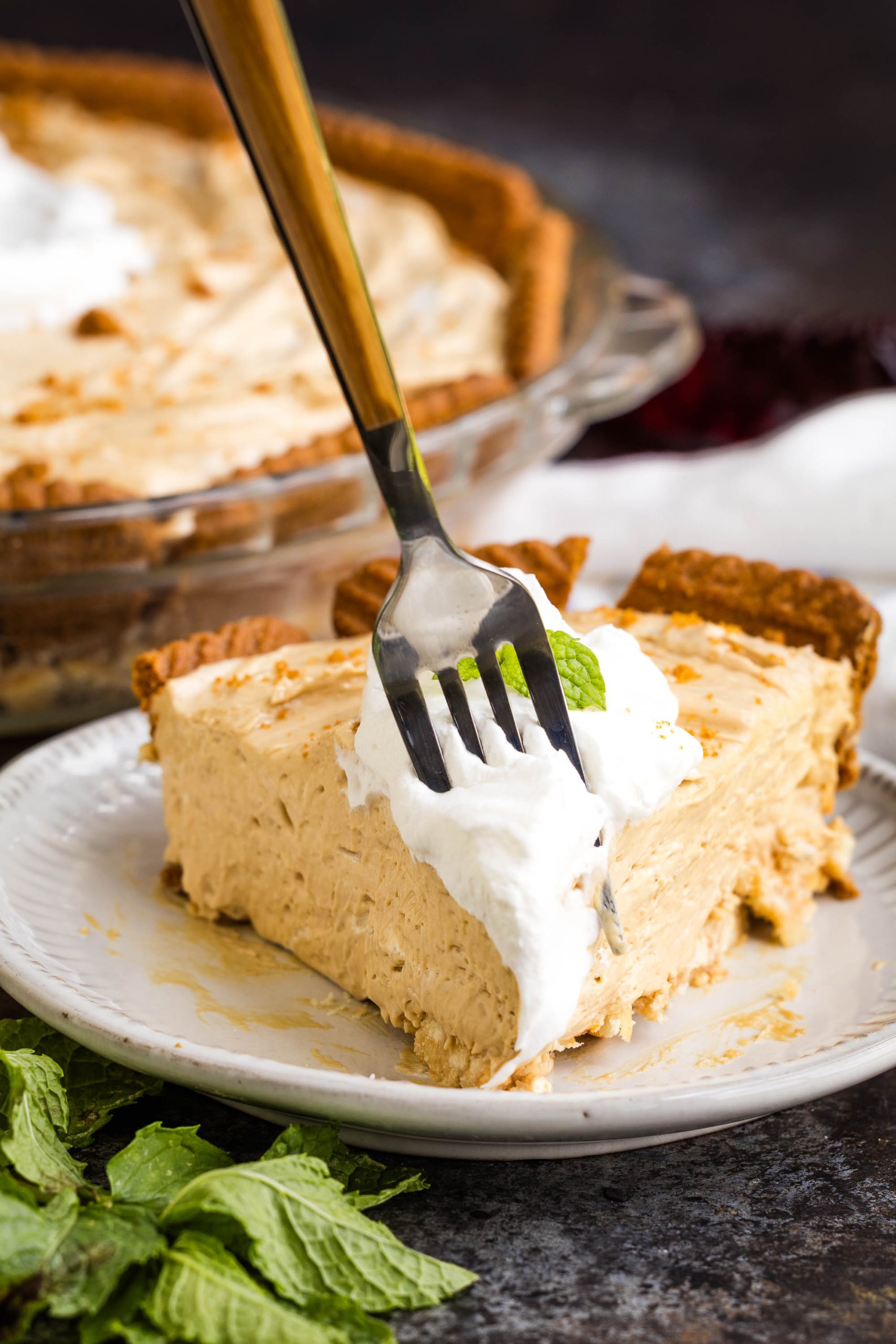 A fork holds a bite of creamy Cookie Butter pie with whipped topping on a plate, with the rest of the pie in a glass dish in the background. Mint leaves are also visible on the table.