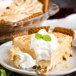 A slice of creamy pie with a graham cracker crust is topped with whipped cream and mint leaves, served on a white plate with a fork.