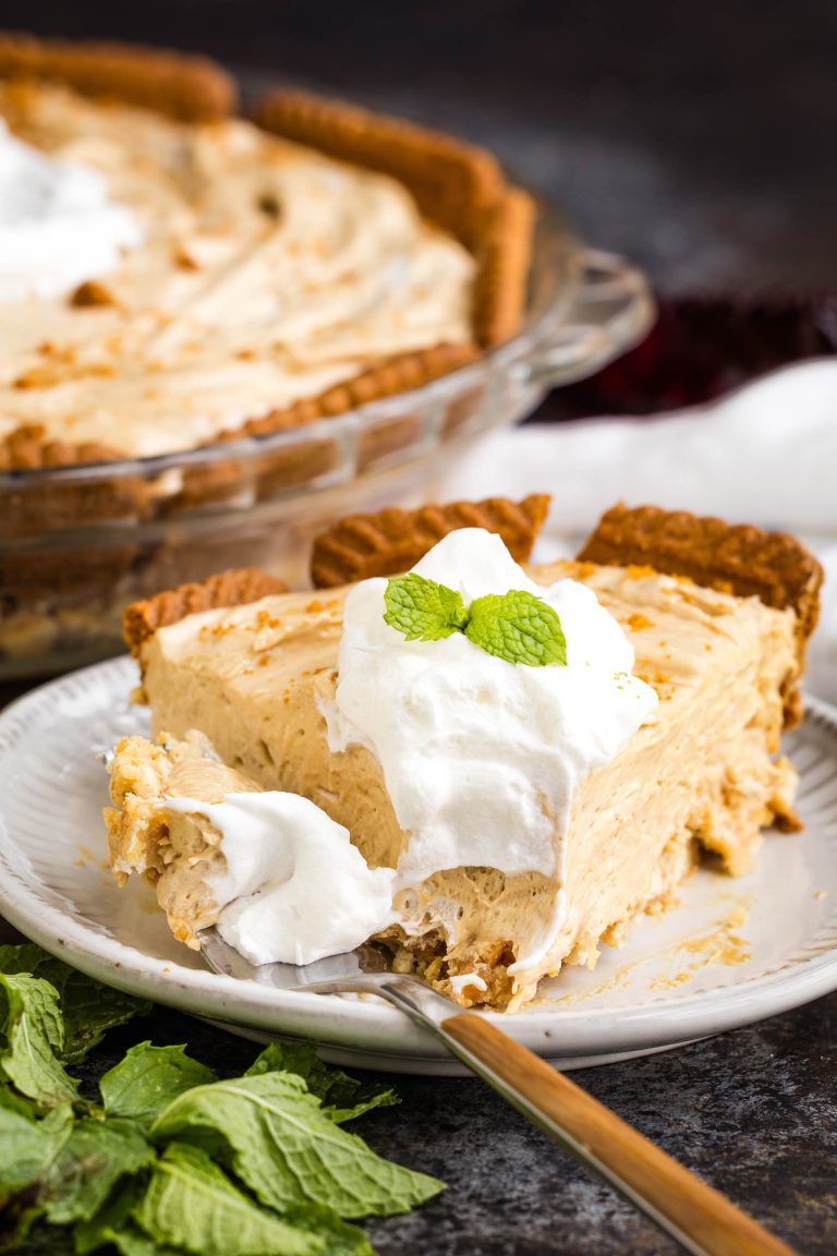 A slice of creamy pie with a graham cracker crust is topped with whipped cream and mint leaves, served on a white plate with a fork.