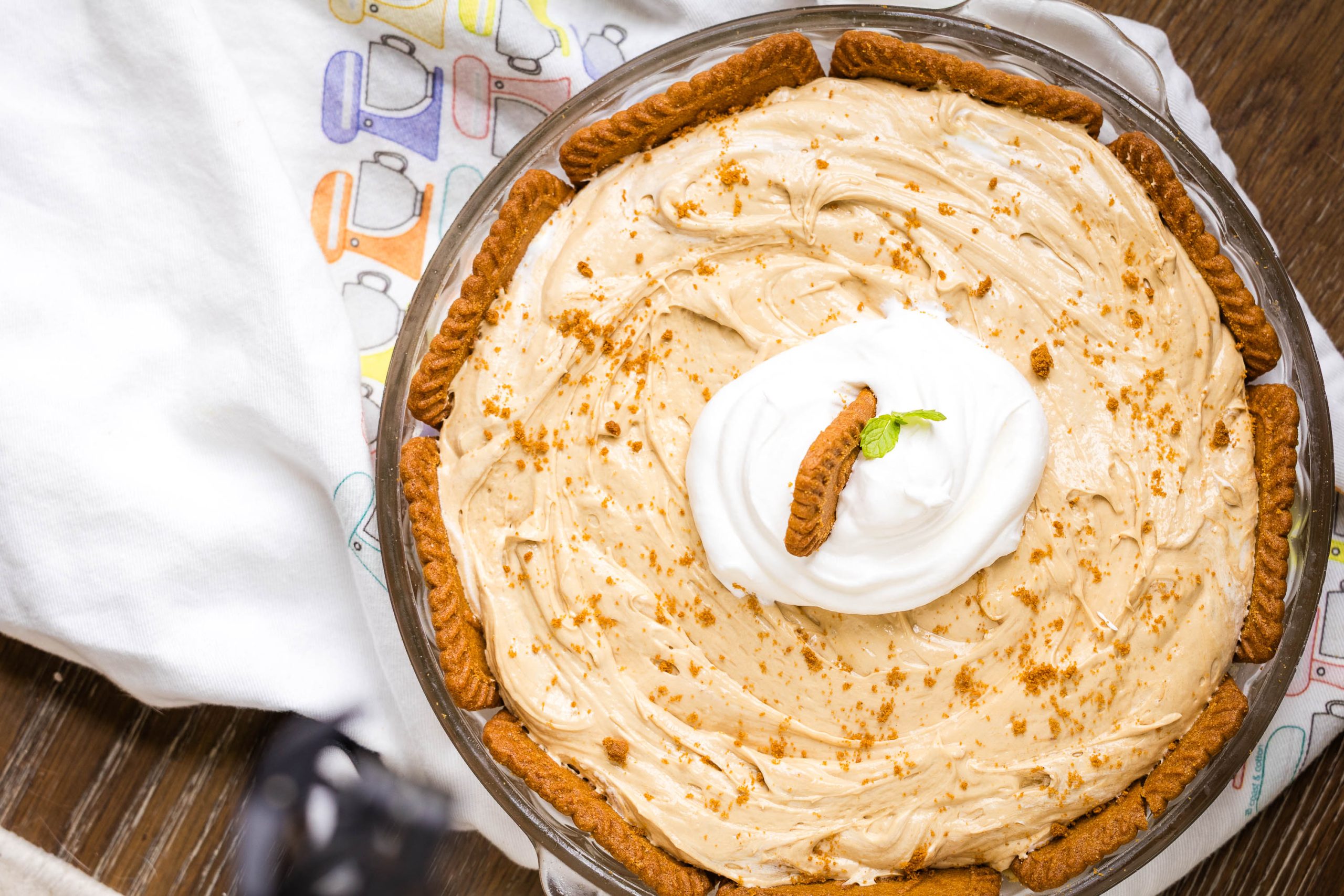A round pie with creamy filling topped with whipped cream, a cookie, and a mint sprig, surrounded by cookies along the crust, on a patterned cloth.