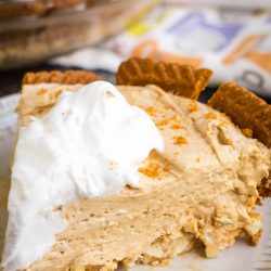 A slice of creamy pie with a graham cracker crust, topped with a dollop of whipped cream, on a white plate.