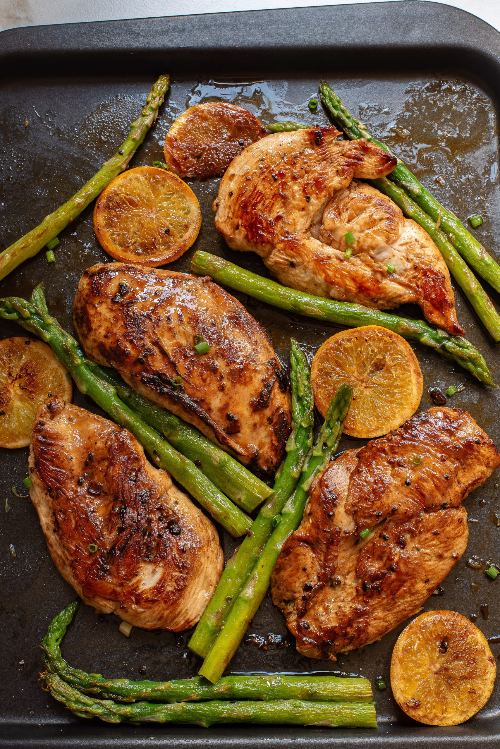 Photo features four cooked chicken breasts, asparagus spears, and round orange slices, all perfectly arranged on a baking sheet for a vibrant and delicious meal.
