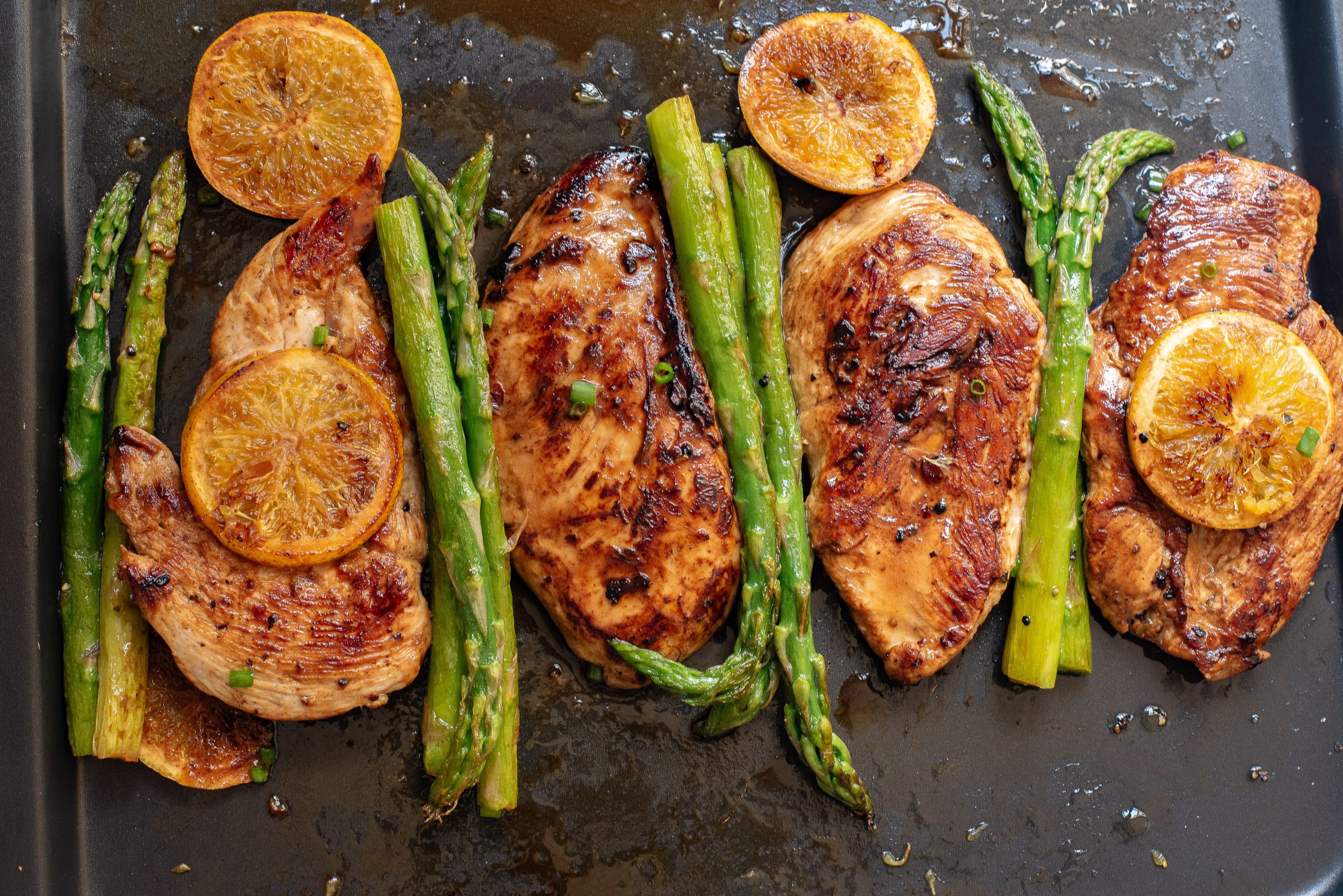 Four cooked chicken breasts garnished with lemon slices and accompanied by asparagus spears on a baking sheet.