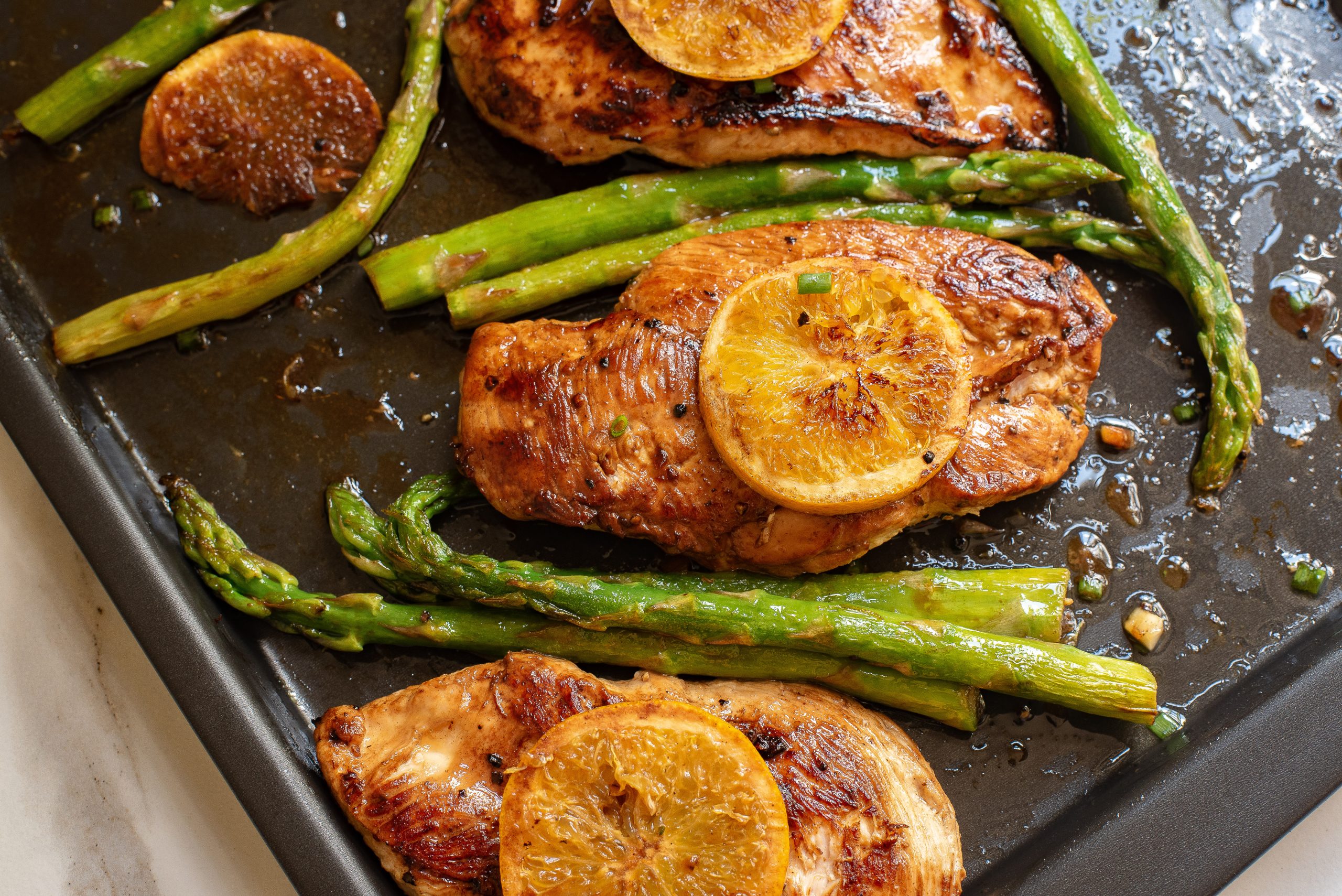Baked chicken breasts topped with lemon slices and served with asparagus spears on a dark baking tray.