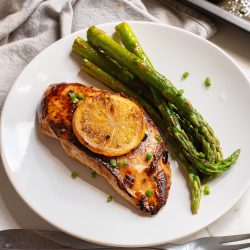 Grilled chicken breast topped with a lemon slice, served alongside roasted asparagus, offers a fresh twist reminiscent of Sheet Pan Orange Chicken and Asparagus on a simple white plate next to a fork and knife.