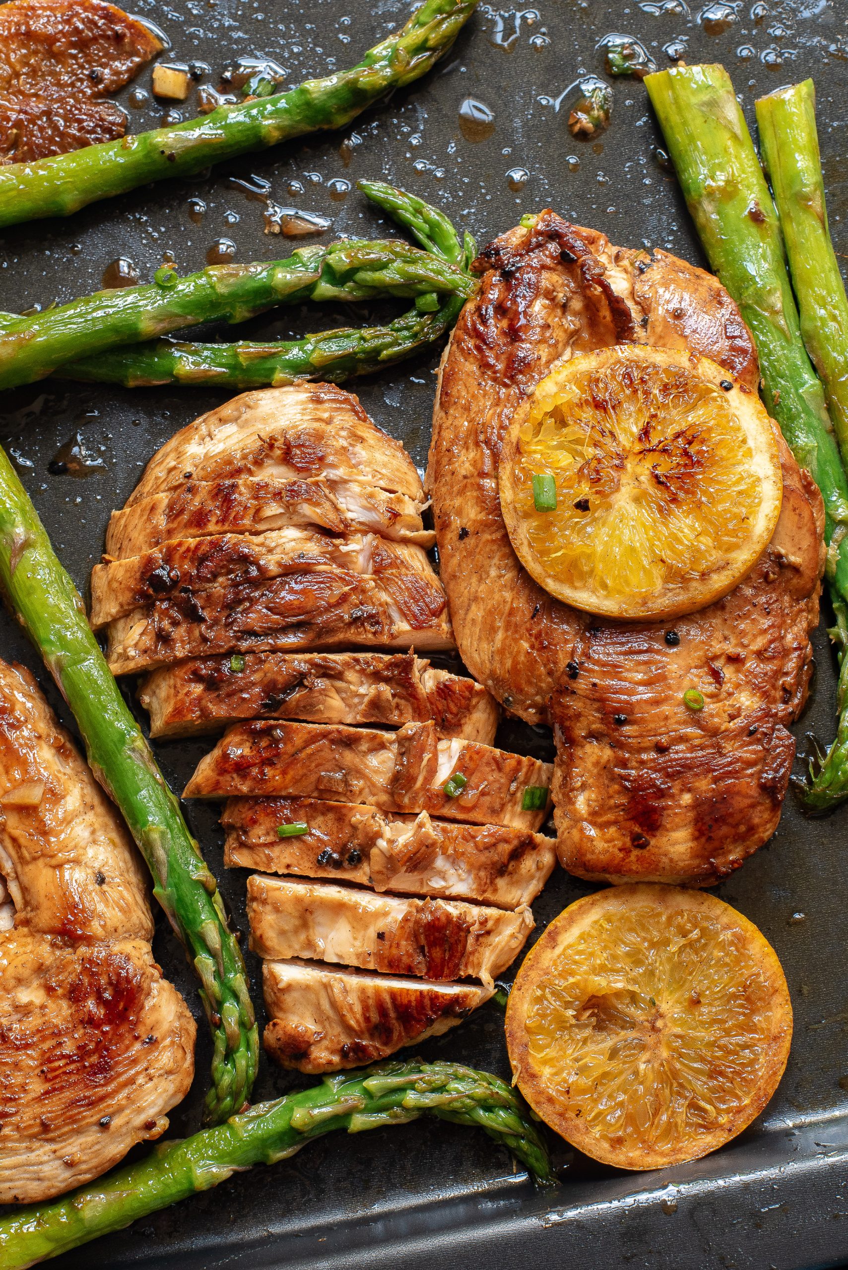 Sheet Pan Orange Chicken and Asparagus features cooked chicken breasts—one sliced, one whole—topped with orange slices and served alongside roasted asparagus spears on a baking tray.